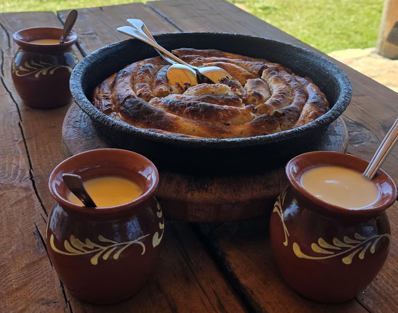 A round burek in a black pan, clay pots of sour milk beside it