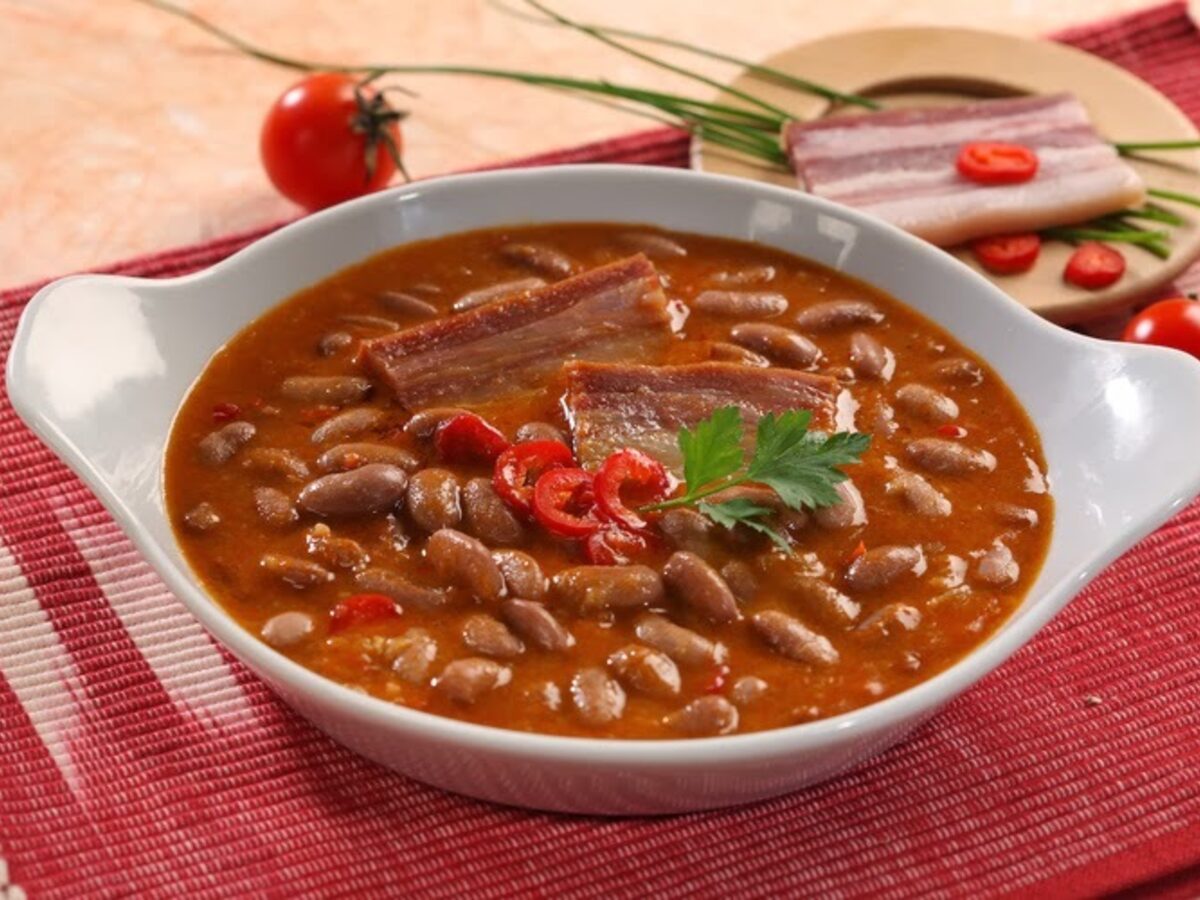 An oval dish of brown bean stew with strips of smoked meat, red chilli and parsley, on a red cloth