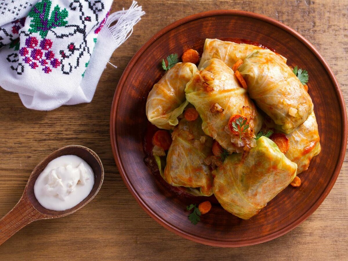 A clay bowl of cabbage rolls with carrots, beside an embroidered cloth and a spoon of sour cream