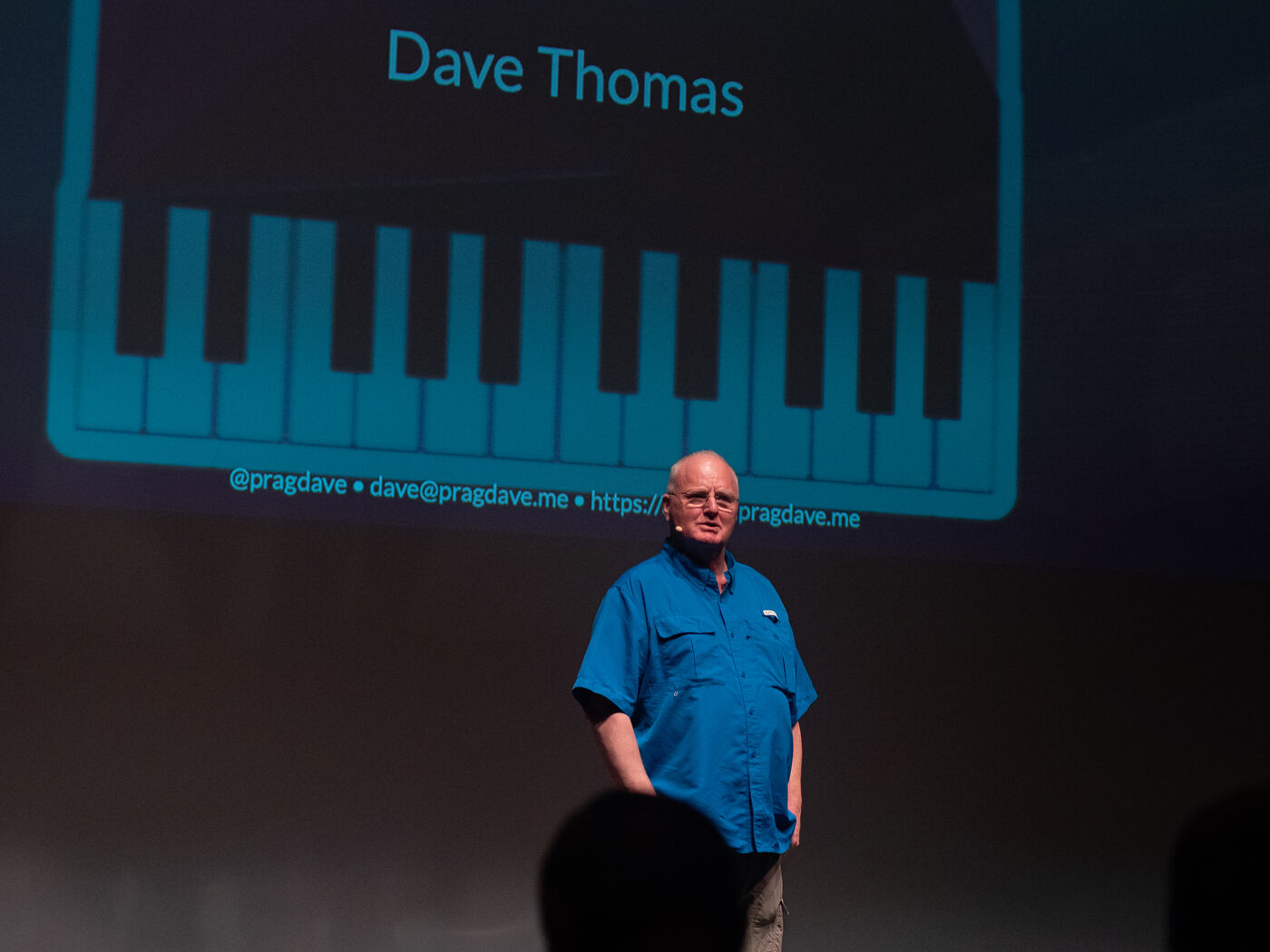 Dave Thomas on stage in a blue shirt, his name on the slide behind him