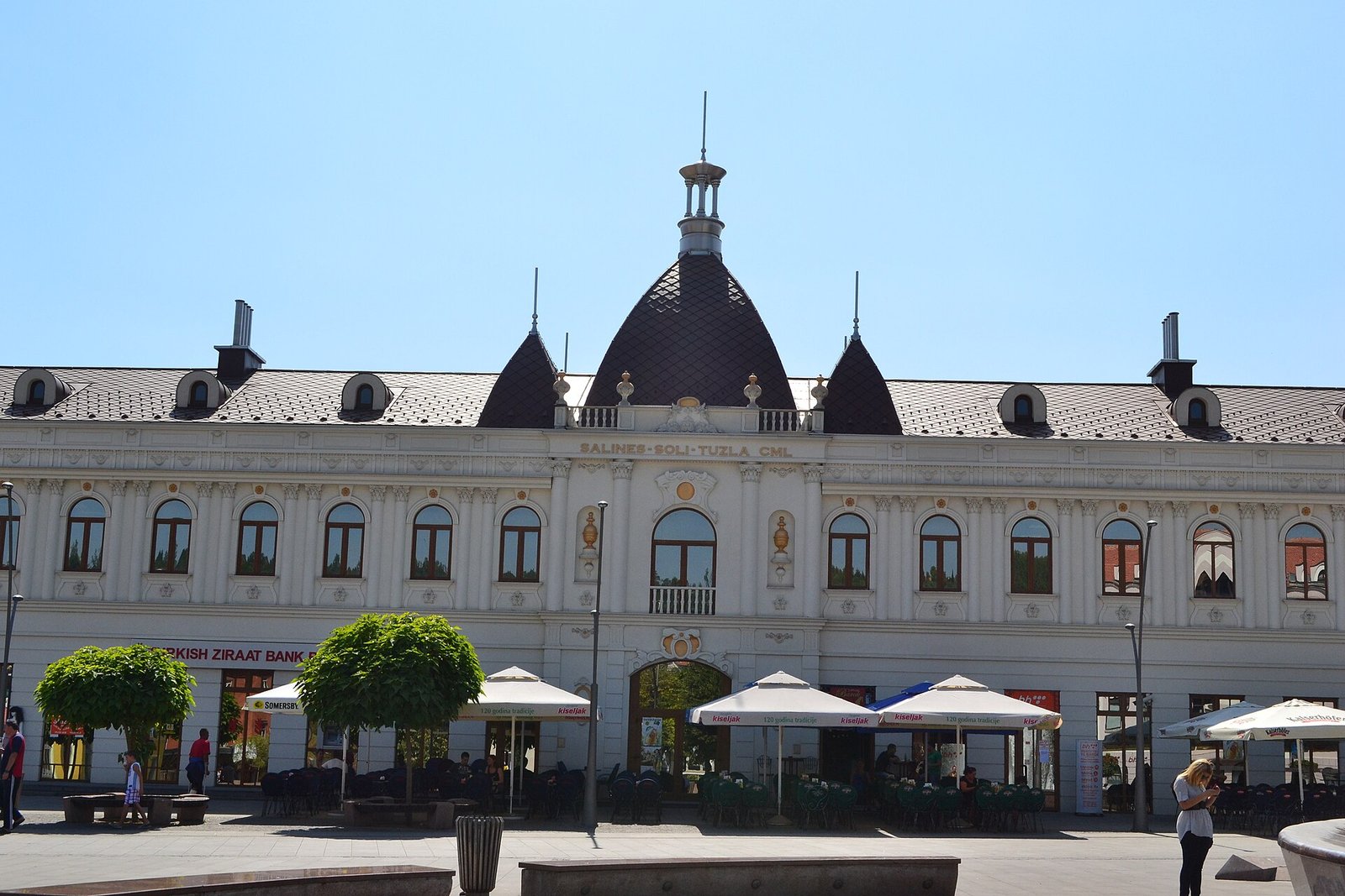 The white neo-Baroque building on Trg slobode with its central dome and two smaller domes, seen head-on in sunshine, with cafe parasols and green terrace chairs along the ground floor