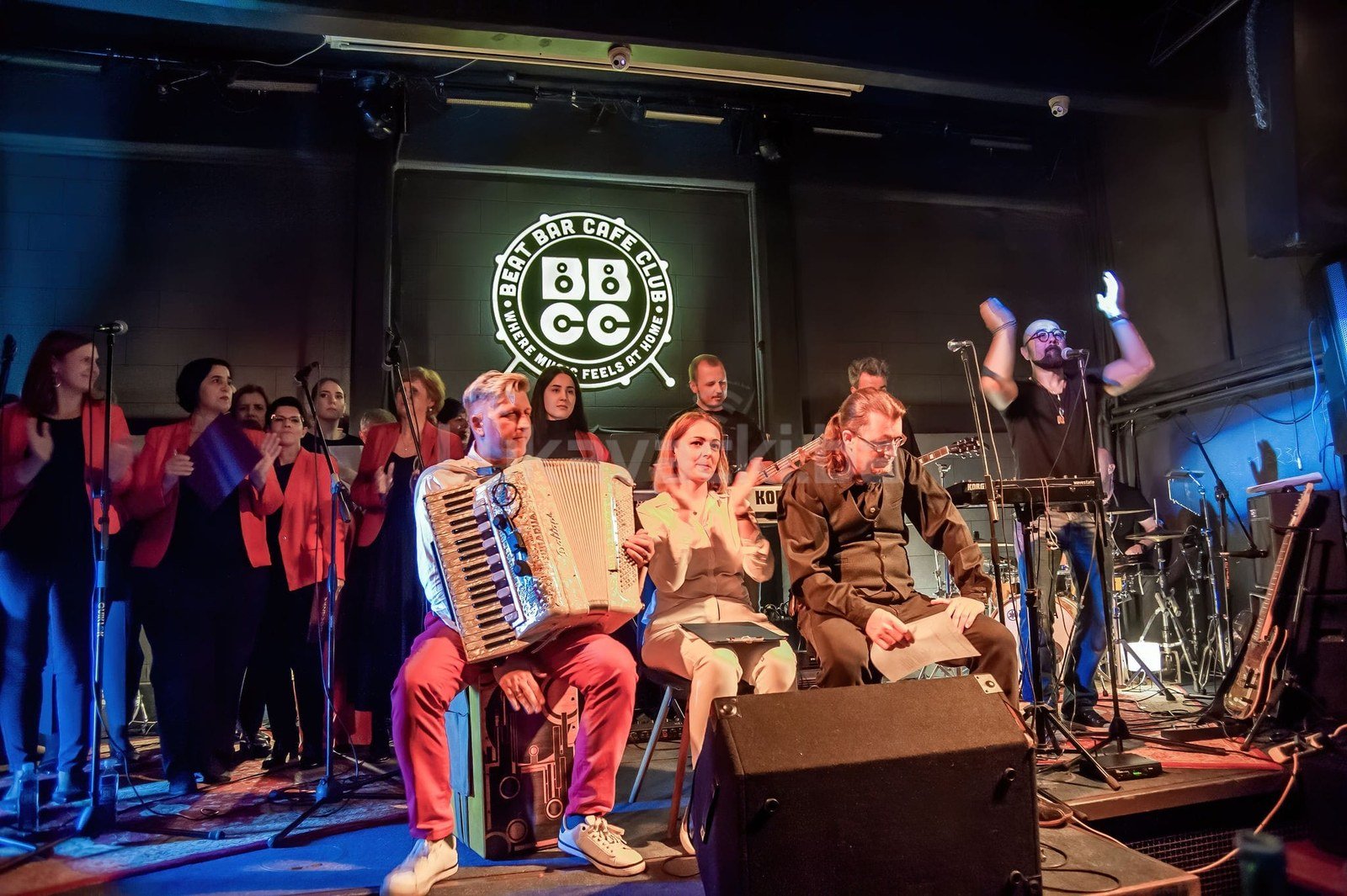 The club's stage under the BBCC Beat Bar Cafe Club logo during a music and poetry night, with a choir, an accordionist and other performers