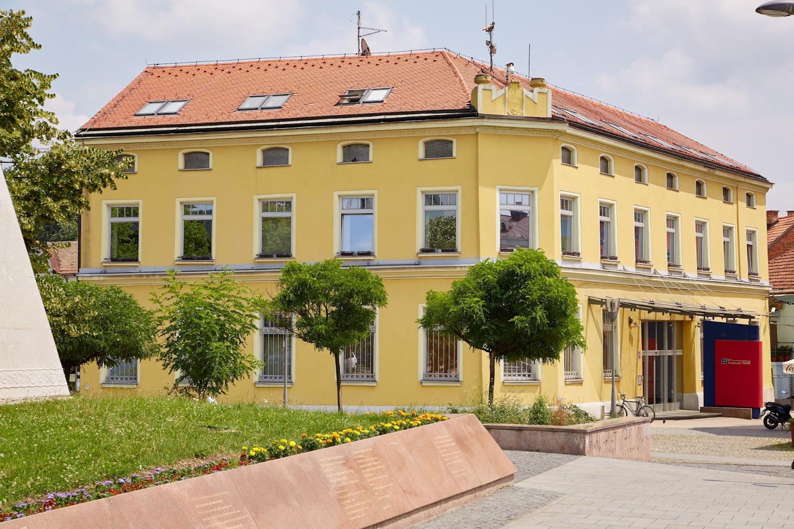 The yellow corner building of BIT Centar with a red tiled roof, small trees and a flower bed in front, and a red and blue BIT Technology Park sign beside the glass entrance