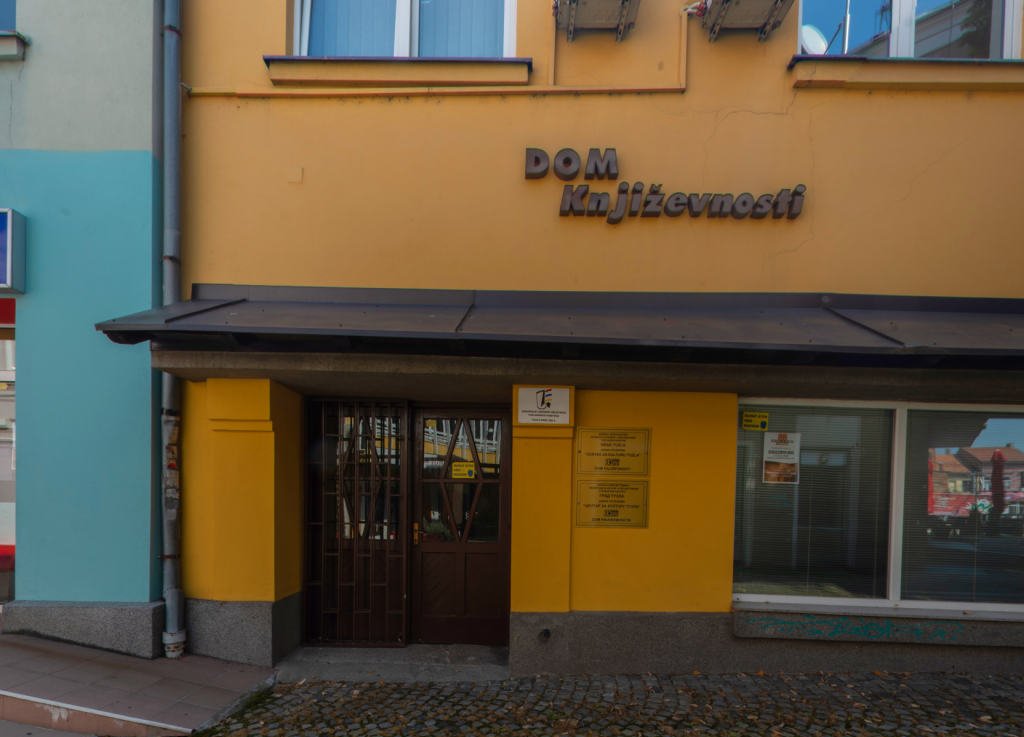 The yellow front of Dom književnosti with its lettered sign, entrance door and windows, seen across the cobbled square in daylight with nobody in frame