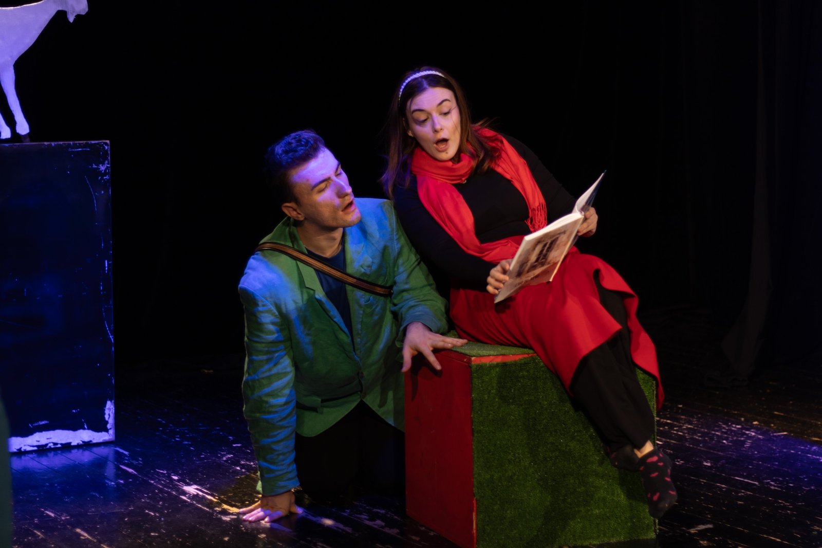 Two actors in costume on a dark stage at Dom mladih, one reading from a book