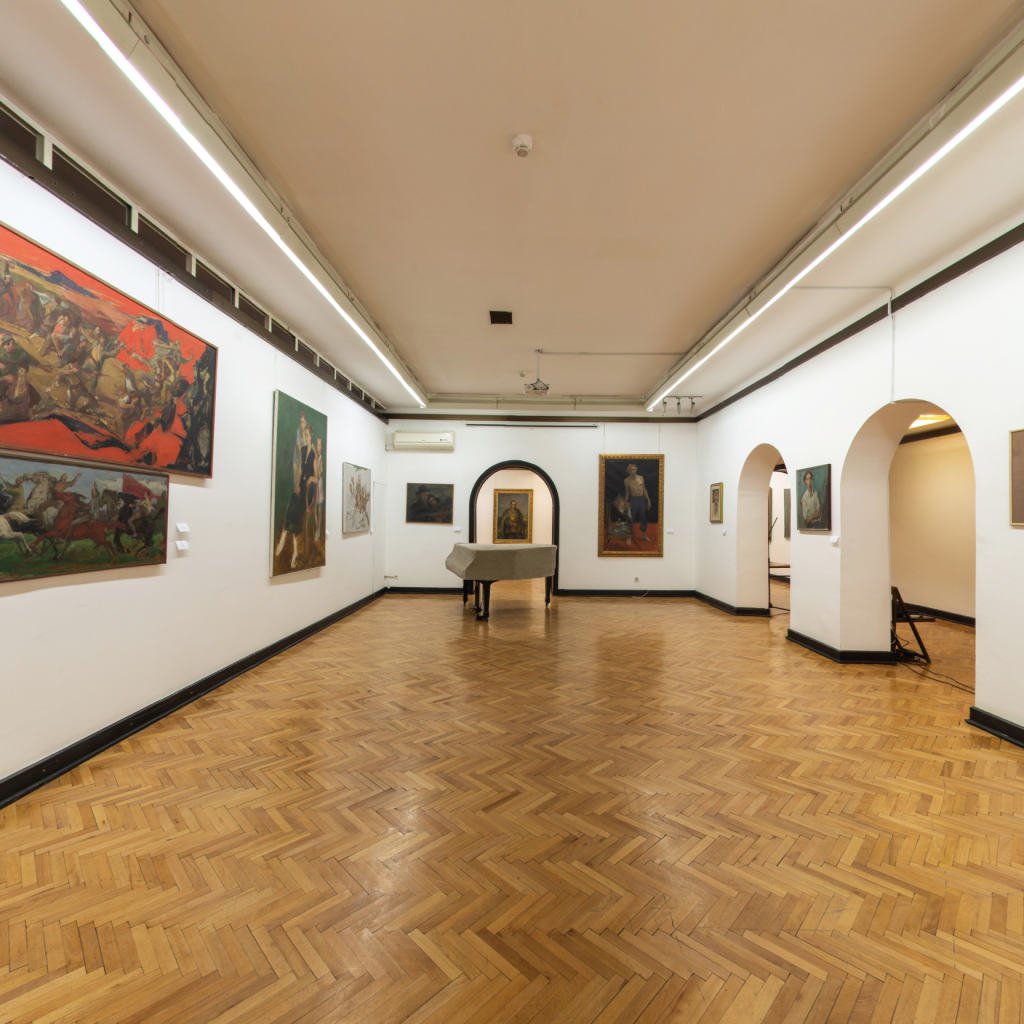 A long, empty, white-walled exhibition hall with paintings hung in a row and a polished herringbone parquet floor
