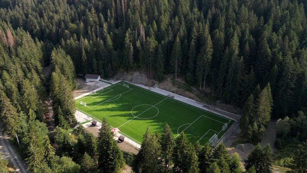 An aerial view of an artificial-grass football pitch set in conifer forest, which the article places at the Muška voda complex