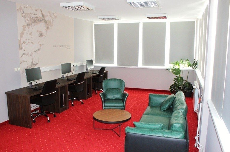 The Student HUB room in 2019, with a red carpet, a row of computer desks along the windows, green armchairs and a green sofa around a round table