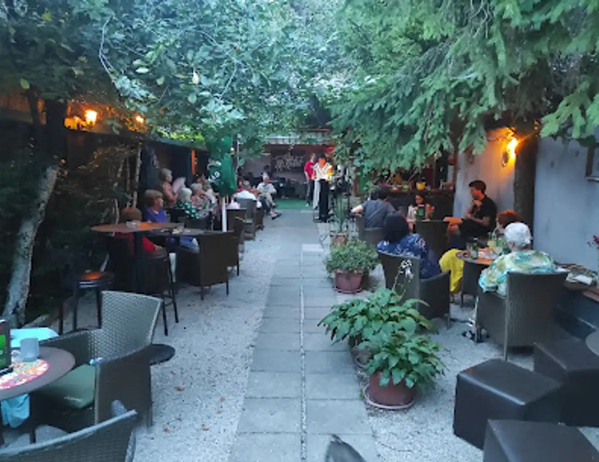 A leafy garden courtyard at dusk with wicker chairs, small tables and potted plants, guests seated along both sides and a performer at the far end