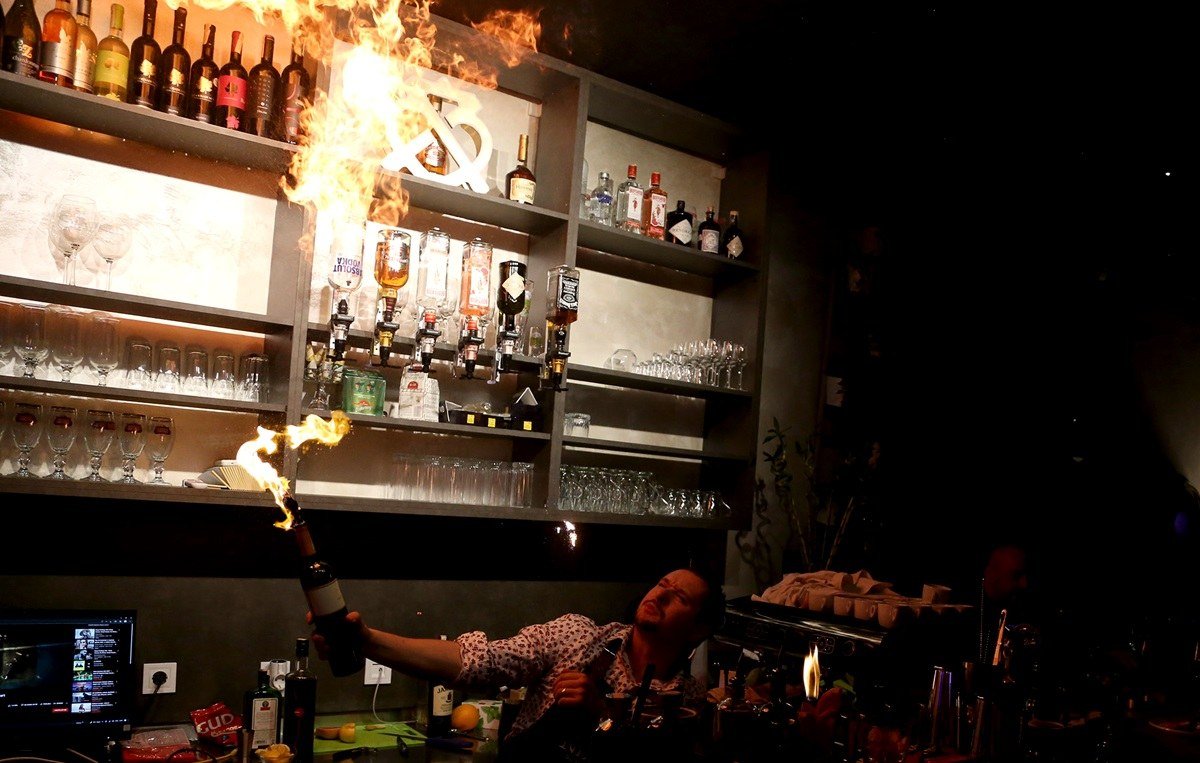 The back bar of Hugo's, shelves of lit bottles, with flames from a bartender's fire show on the opening night