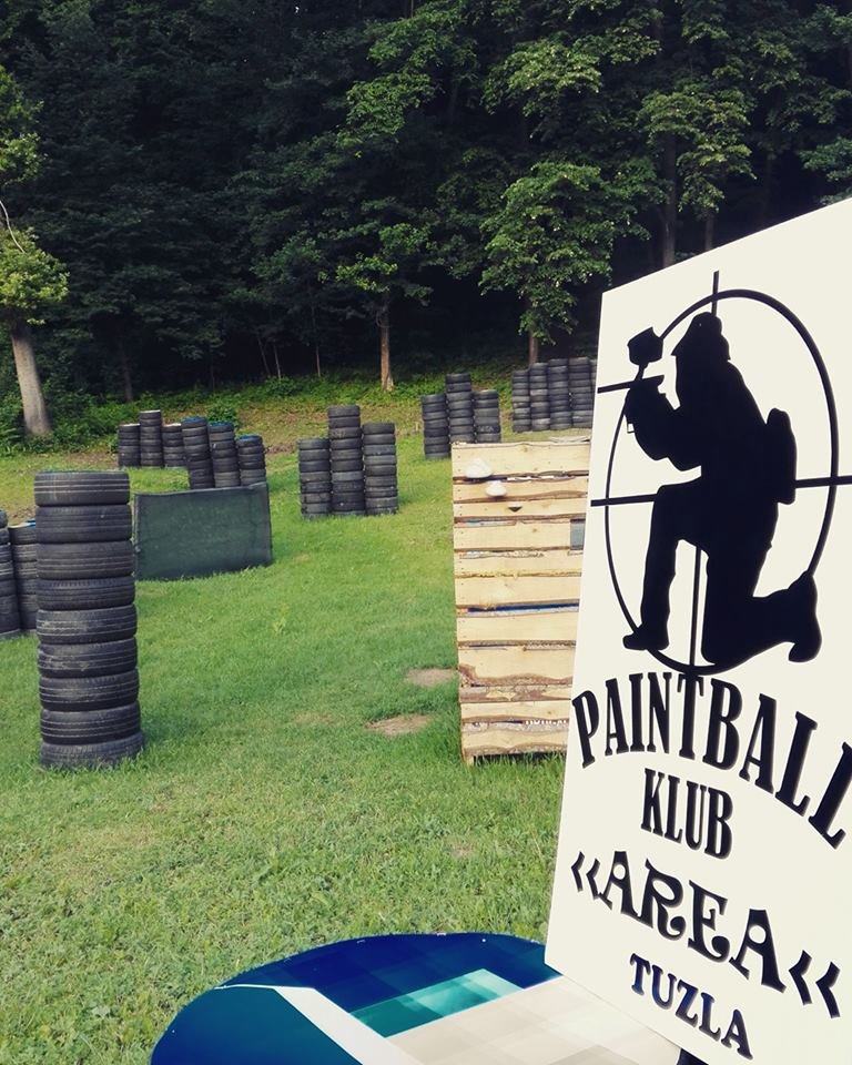 The club's paintball field: tyre stacks and wooden barricades on grass by a forest, with the club's sign in front