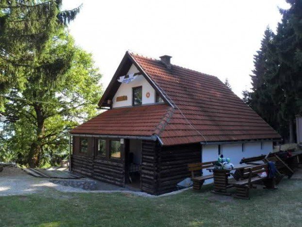 The Javorje mountain lodge in summer, a timber-and-white house with a steep red tile roof, benches and tables in front, among trees