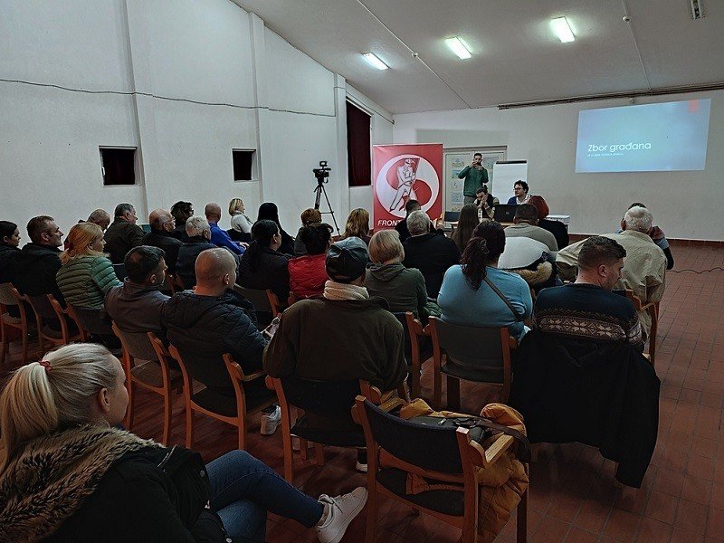 A citizens' meeting in the house's large hall in November 2022, with rows of seated residents facing a projected slide and a speakers' table